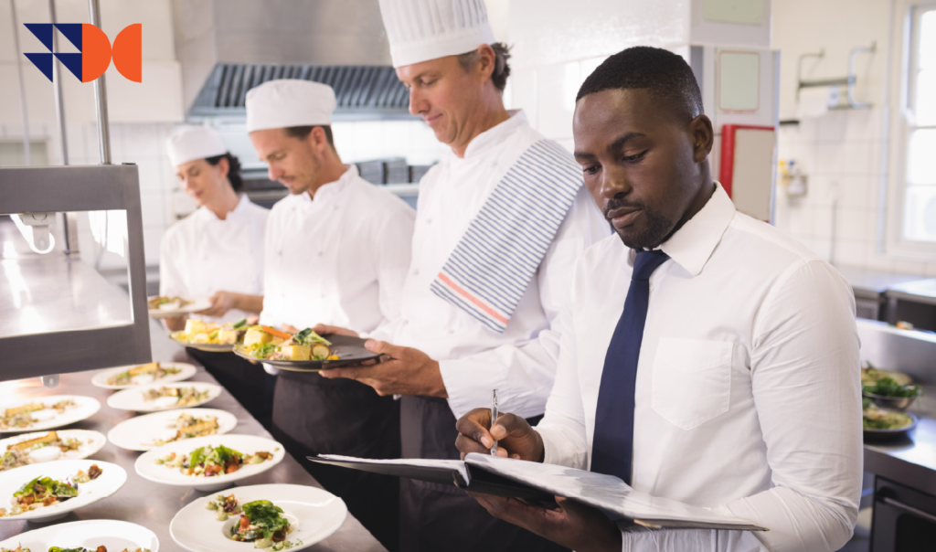 Restaurant manager reviewing records while chefs plate food, representing employer compliance with the extended Fast Food, Restaurant and Catering Bargaining Council agreement