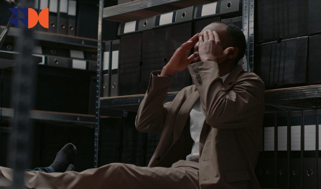 Distressed employee sitting on the floor of an office archive with his hands over his face, illustrating depression and workplace absence